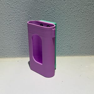 Op de afbeelding: Een 3D-geprinte behuizing in lavendel met een rechthoekige vorm en een gebogen uitsparing. Het heeft een groene achterkant en twee kleine zwarte cirkelvormige elementen aan de bovenkant. Het item is waarschijnlijk ontworpen om een klein elektronisch apparaat te bevatten.