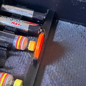 Könnte beinhalten: Ein schwarzer Kunststoff-Organizer mit mehreren schwarzen AA-Batterien und kleinen orangefarbenen und gelben Objekten. Die Batterien haben rote, gelbe und orangefarbene Bänder. Der Organizer hat eine strukturierte Oberfläche.