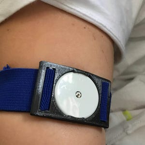 Op de afbeelding: Een zwart-blauwe hartslagmeter met een witte sensor. De monitor wordt op de bovenarm gedragen.