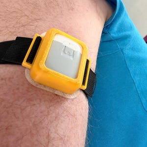 Op de afbeelding: Een medisch hulpmiddel dat met een zwarte band aan de arm is bevestigd. Het apparaat heeft een gele behuizing en een grijs en wit midden. Het apparaat is bevestigd aan een beige zelfklevende pad.