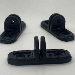 Op de afbeelding: Drie zwarte, 3D-geprinte camerabevestigingen van plastic met elk twee gaten. De bevestigingen zijn ontworpen om aan een oppervlak te worden bevestigd en hebben een gleuf voor het plaatsen van een camera.