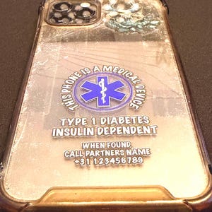 Op de afbeelding: Transparante telefoonhoes met gouden rand. De tekst "THIS PHONE IS A MEDICAL DEVICE" en "TYPE 1 DIABETES INSULIN DEPENDENT" is zichtbaar. Er staat ook een telefoonnummer op.