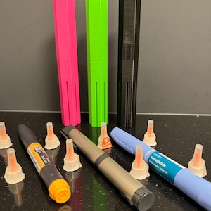 May include: A close-up of various medical supplies, including three colorful pens, two insulin pens, and several needle caps. The pens are black, green, and pink. The insulin pens are silver and blue. The needle caps are white with orange tips. The blue pen has the text "Novo Nordisk semaglutide subcutaneous gebruik" printed on it.