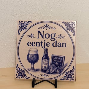 Op de afbeelding: Een vierkante keramische tegel met een blauw-wit ontwerp en de tekst Nog eentje dan. De illustratie bevat een bierglas, een fles met het label Speciaal Bier en een bordje met de tekst Lekker Speciaal, omringd door florale motieven.