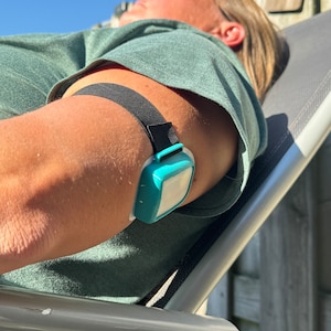 Op de afbeelding: Een turquoiseblauw medisch apparaat met een wit gezicht is bevestigd aan de arm van een persoon met een zwarte band. Het apparaat wordt op de bovenarm gedragen.