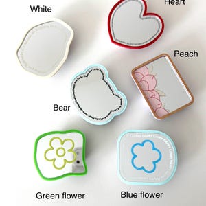 May include: A collection of six small handheld mirrors in various shapes and colors. The set includes a white irregular mirror, a red heart-shaped mirror, a peach-themed rectangular mirror, a bear-shaped mirror, a green flower mirror, and a blue flower mirror with text.