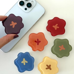 May include: A collection of colorful flower-shaped phone grips in shades of red, orange, yellow, green, blue, and purple. Each grip features a contrasting cross design in the center. One grip is attached to a silver smartphone.