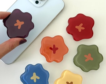 May include: A collection of colorful flower-shaped phone grips in shades of red, orange, yellow, green, blue, and purple. Each grip features a contrasting cross design in the center. One grip is attached to a silver smartphone.