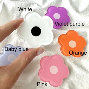 Op de afbeelding: Vijf bloemvormige telefoonbeugels in verschillende kleuren: wit, violetpaars, oranje, roze en babyblauw. Elke beugel heeft een cirkel in het midden.