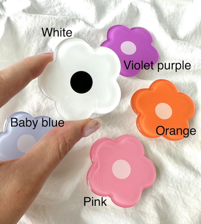 Op de afbeelding: Vijf bloemvormige telefoonbeugels in verschillende kleuren: wit, violetpaars, oranje, roze en babyblauw. Elke beugel heeft een cirkel in het midden.