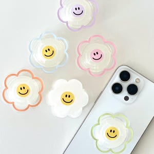 May include: Six colorful phone grips shaped like flowers with a smiley face in the center. The grips are made of clear plastic with a colored outline. One grip is on a white iPhone.