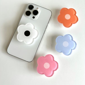 Op de afbeelding: Vier telefoonbeugels in de vorm van een bloem. De beugels zijn wit, roze, blauw en oranje. De witte beugel heeft een zwart midden, de roze beugel heeft een wit midden, de blauwe beugel heeft een wit midden en de oranje beugel heeft een wit midden.