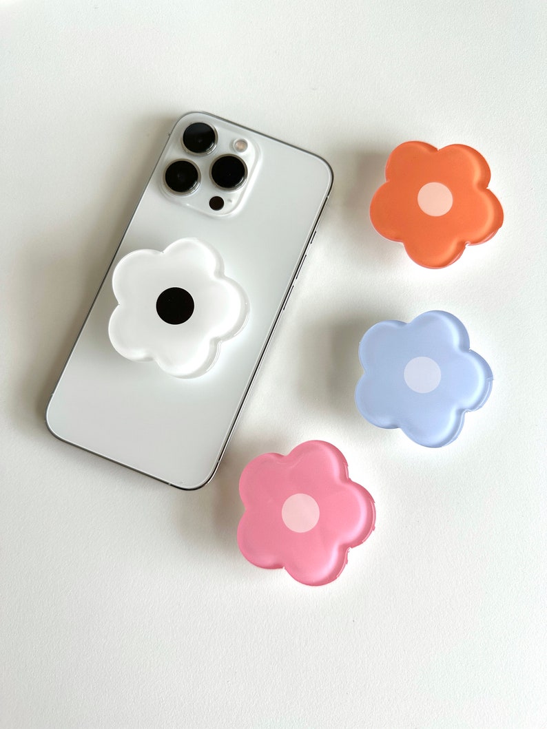 Op de afbeelding: Vier telefoonbeugels in de vorm van een bloem. De beugels zijn wit, roze, blauw en oranje. De witte beugel heeft een zwart midden, de roze beugel heeft een wit midden, de blauwe beugel heeft een wit midden en de oranje beugel heeft een wit midden.