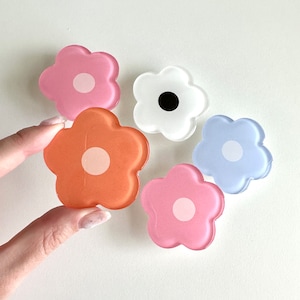 Op de afbeelding: Vijf telefoonbeugels in de vorm van bloemen van hars in verschillende kleuren, waaronder roze, oranje, wit en blauw. Elke beugel heeft een witte cirkel in het midden.