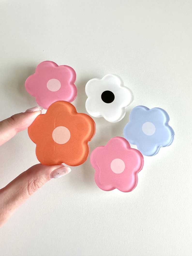 Op de afbeelding: Vijf telefoonbeugels in de vorm van bloemen van hars in verschillende kleuren, waaronder roze, oranje, wit en blauw. Elke beugel heeft een witte cirkel in het midden.