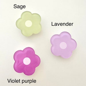 Op de afbeelding: Drie haarclips in de vorm van bloemen gemaakt van hars in de kleuren saliegroen, lavendel en violet.