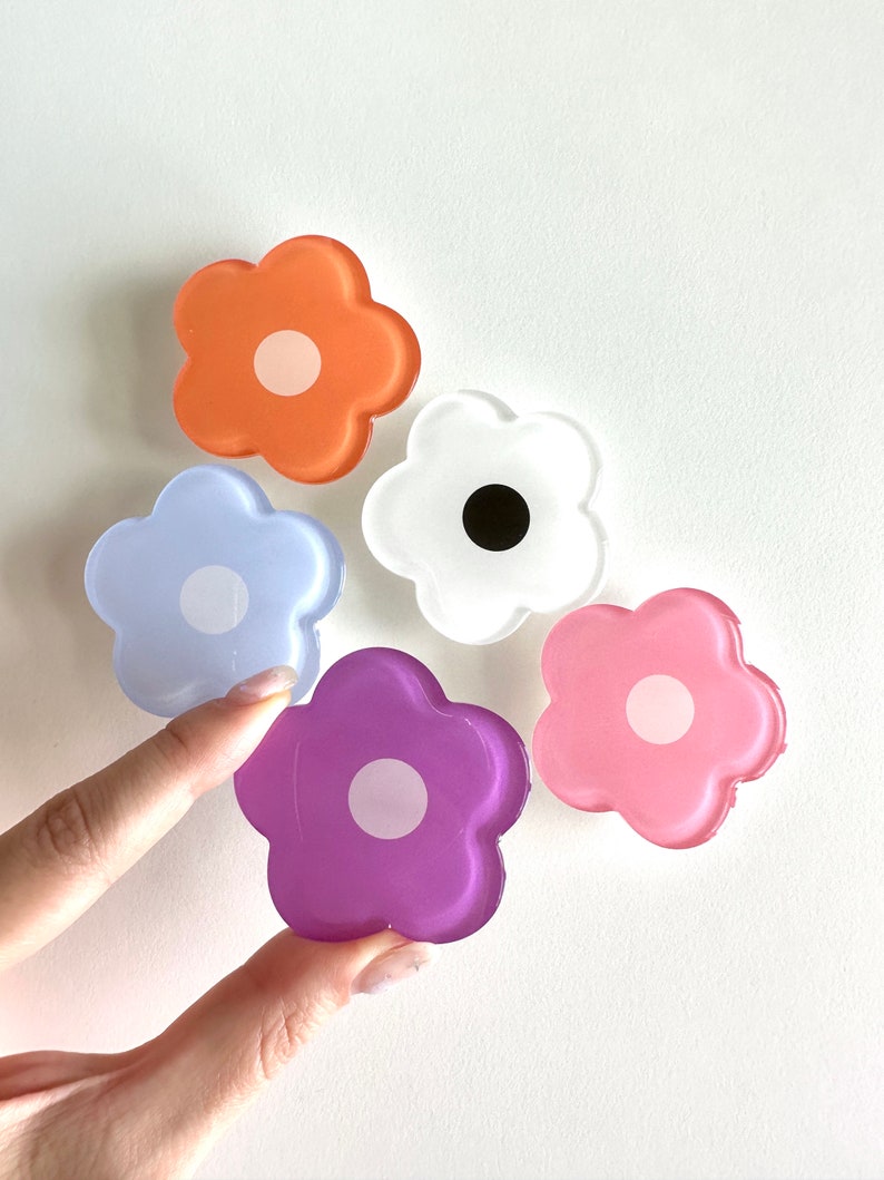 Op de afbeelding: Vijf kleurrijke, bloemvormige telefoonbeugels van hars met witte centra. De bloemen zijn in tinten oranje, wit, lichtblauw, roze en paars.
