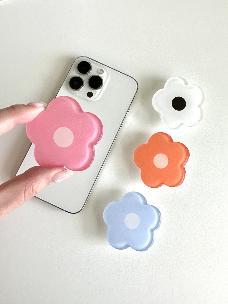 Op de afbeelding: Vier telefoonbeugels in de vorm van een bloem. De beugels zijn roze, oranje, lichtblauw en wit. De witte beugel heeft een zwart midden, terwijl de andere drie een wit midden hebben.