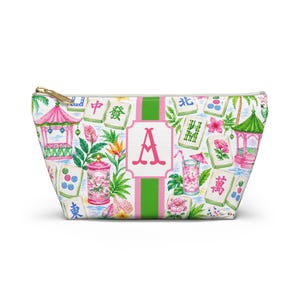 May include: A white zipped pouch with a colourful pattern featuring pink pagodas, mahjong tiles, tropical flowers, and a pink letter "A" on a green and white striped background. The pouch has a gold zip.