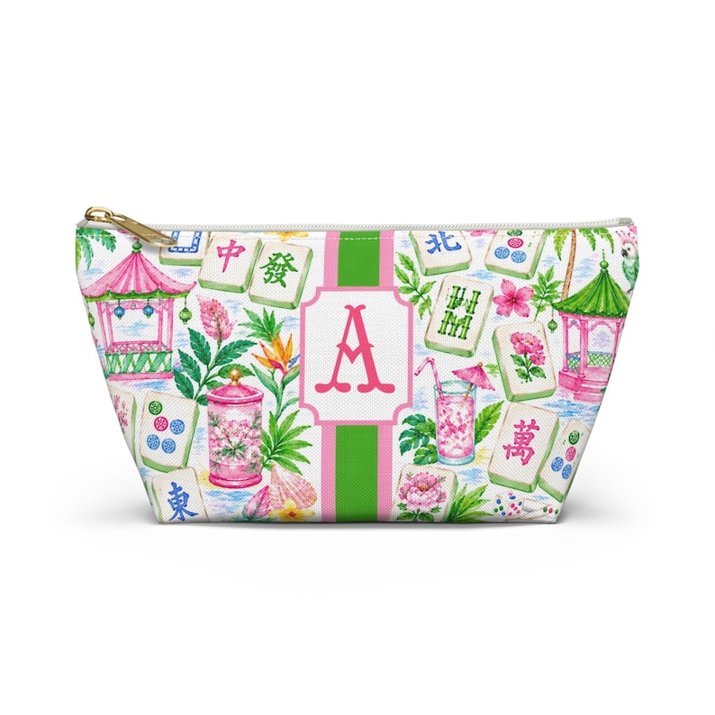May include: A white zipped pouch with a colourful pattern featuring pink pagodas, mahjong tiles, tropical flowers, and a pink letter "A" on a green and white striped background. The pouch has a gold zip.