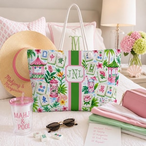 May include: A patterned tote bag with a Mahjong tile design, a straw hat, and sunglasses. The bag has a pink and green stripe and the initials "JNL". A tumbler with "MAHJ & POOL" text is also visible.