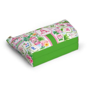 May include: A rectangular cosmetic bag with a green base and a patterned top. The top features a floral and tile design with pink and green accents. A pink stripe with the letter "A" is centered on the top. A gold zip is visible.