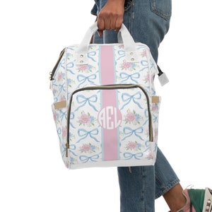 May include: A white backpack with a floral and bow pattern, a pink stripe, and a monogram. The bag has a top handle, side pockets, and a front pocket. The floral pattern includes pink and blue flowers.