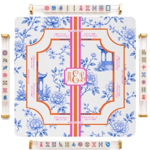 May include: A square mahjong mat featuring a blue and white floral toile pattern with orange and pink accents. The center displays the monogram AEL. Four clear acrylic tile racks with gold-tone ends are positioned around the mat, each holding mahjong tiles.