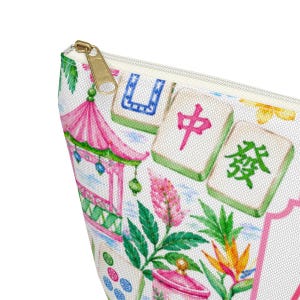 May include: A white cosmetic bag with a gold zip, featuring a colourful print of a pink pagoda, green leaves, and Mahjong tiles. The bag has a rectangular shape and a cream-coloured interior.