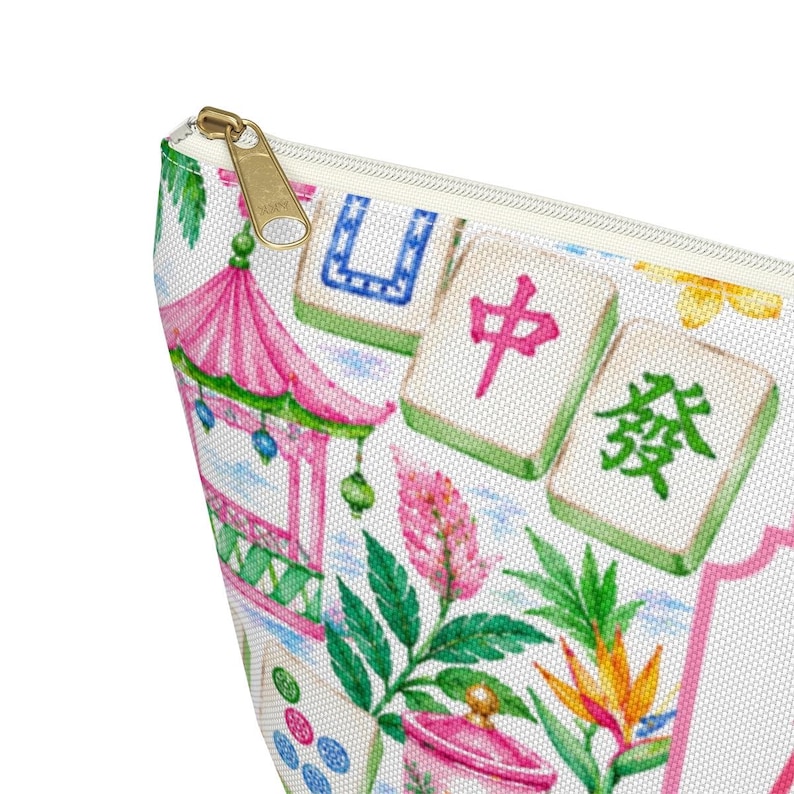 May include: A white cosmetic bag with a gold zip, featuring a colourful print of a pink pagoda, green leaves, and Mahjong tiles. The bag has a rectangular shape and a cream-coloured interior.