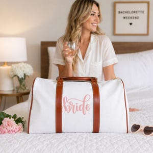 May include: A white duffel bag with brown leather accents and handles. The word "bride" is written in pink script across the front. The bag is on a white bed, with a woman in the background. A sign that says "Bachelorette Weekend" is on the wall.
