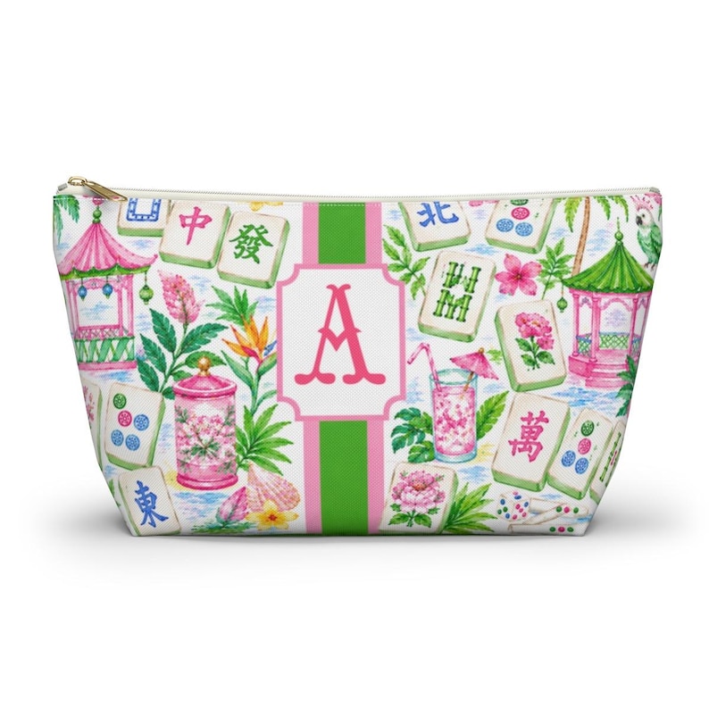 May include: A zipped pouch with a vibrant print of pink pagodas, tropical flowers, and Mahjong tiles. The design includes a large pink letter "A" on a white and green striped background. The pouch has a gold zip.