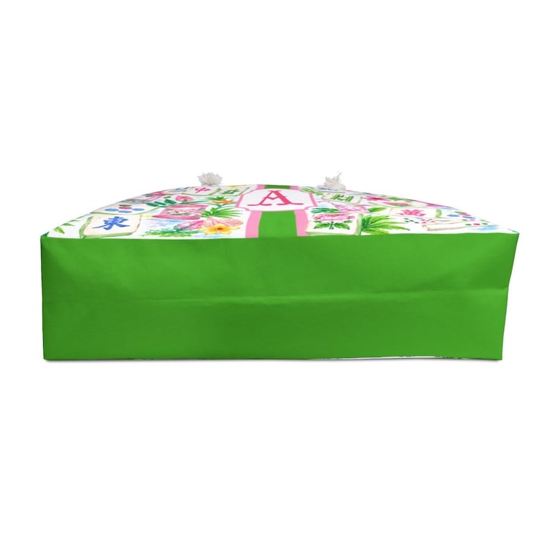 May include: A tote bag with a bright green base and a white top featuring a floral pattern with pink accents. A pink stripe with the letter "A" is centered on the top. The bag has white rope handles.