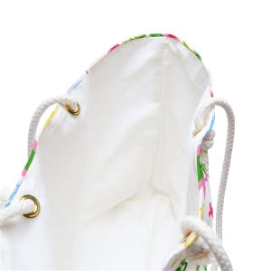 May include: White tote bag with a floral pattern in pink, green, and yellow. The bag has a white interior and rope handles attached with gold eyelets. The bag is designed for carrying items.