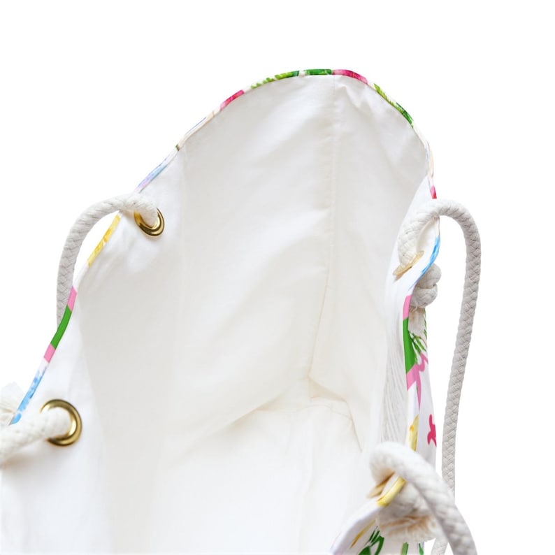 May include: White tote bag with a floral pattern in pink, green, and yellow. The bag has a white interior and rope handles attached with gold eyelets. The bag is designed for carrying items.