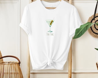 Könnte beinhalten: Weißes T-Shirt mit einem grünen Martini-Glas mit Oliven und dem aufgedruckten Text "Tini Time".