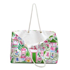 May include: White tote bag with a colourful print featuring pink pagodas, floral arrangements, and Mahjong tiles. The bag has a white rope handle and a green and pink stripe down the centre.