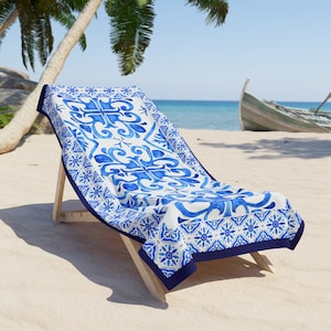 May include: A rectangular beach towel featuring a white base with a repeating blue watercolor-style floral and geometric tile pattern. The towel has a solid dark blue border and is draped over a wooden lounge chair on a sandy beach.
