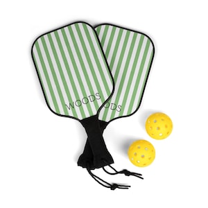 May include: Two green and white striped pickleball paddles with black handles and the word "WOODS" printed on one paddle. Two yellow pickleball balls are included.