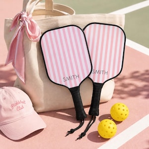Könnte beinhalten: Zwei Pickleball-Schläger mit rosa und weißen Streifen und dem Aufdruck "SMITH". Die Schläger haben schwarze Griffe und liegen neben zwei gelben Pickleballs, einer rosa Kappe mit dem Text "Pickleball Club" und einer beigen Tragetasche mit einem rosa Schal.