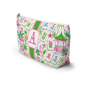 May include: A white cosmetic bag with a gold zip, decorated with a vibrant pattern of Mahjong tiles, tropical flowers, and a pink and green striped design. The letter "A" is displayed in the centre.