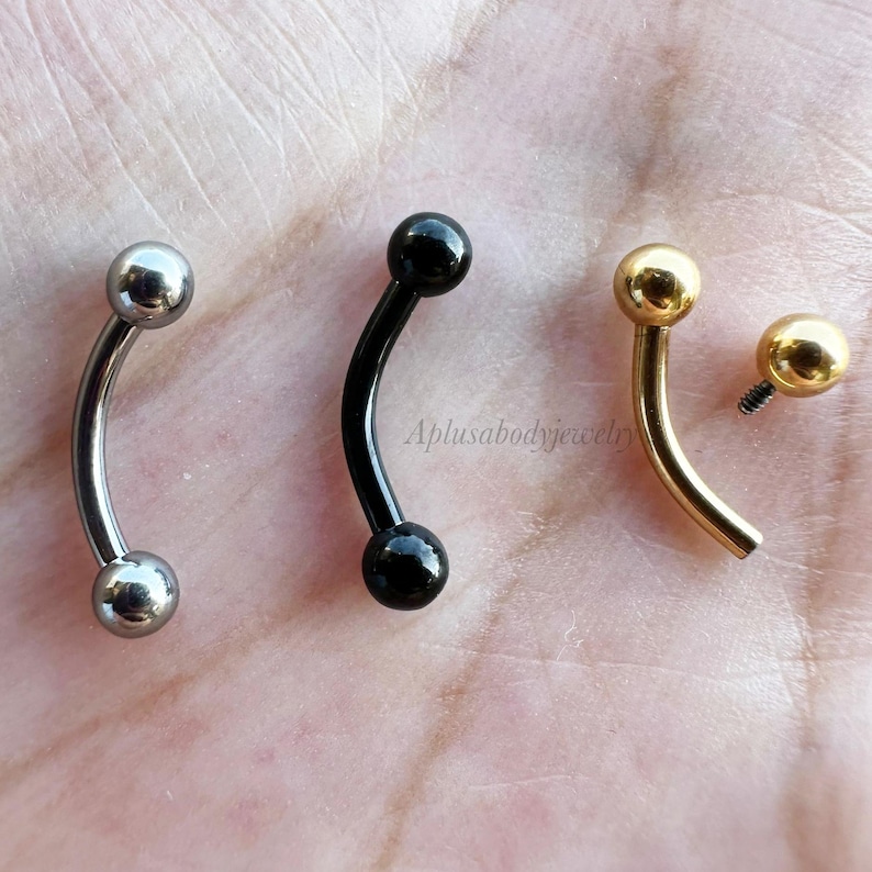 May include: Three curved barbell body jewelry pieces with different colored balls. The first is silver with silver balls, the second is black with black balls, and the third is gold with gold balls. Aplusabodyjewelry is printed on the image.
