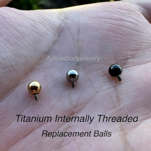 May include: Three titanium internally threaded replacement balls in gold, silver, and black. The balls are round with a threaded post for body jewelry. Text on the image reads "Titanium Internally Threaded Replacement Balls."