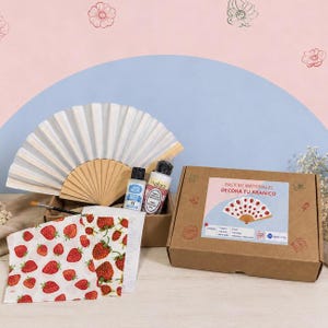 Puede incluir: Un kit de manualidades para decorar un abanico, que incluye un abanico de papel blanco con marco de madera, dos botes de pintura, un pincel, una hoja con estampado de fresas y una caja de cartón marrón con la etiqueta Pack de Materiales.
