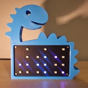 Peut inclure: Lampe LED bleue en forme de dinosaure. La lampe présente un panneau rectangulaire noir avec de nombreuses petites lumières bleues vives. Le dinosaure a un visage amical et un dos crénelé. La lampe est en matériau bleu clair.