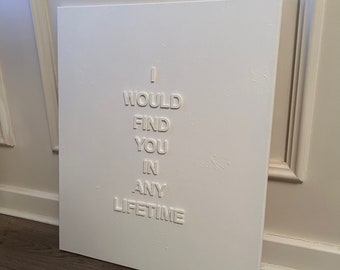 May include: A white rectangular canvas featuring raised, three-dimensional white lettering that spells out the phrase I WOULD FIND YOU IN ANY LIFETIME. The surface of the canvas has a subtle, textured finish, and the letters are centered.