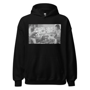 Op de afbeelding: Zwart-witfoto van twee mensen die aan een tafel zitten met een stapel geld voor hen. De persoon links draagt een shirt met lange mouwen en de persoon rechts draagt een shirt met korte mouwen.