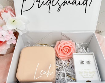 May include: A white box with the text "Will you be my bridesmaid?" on the lid. The box is open and contains a pink jewellery box with the name "Leah" on it, a pink rose, and a small white box with a pair of earrings and the text "I couldn't tie the knot without you".