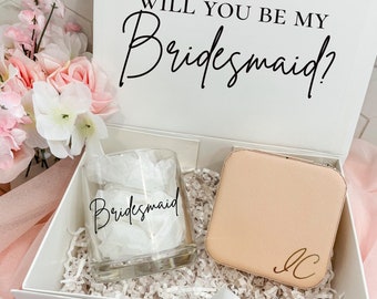 Op de afbeelding: Een witte cadeaudoos met een wit deksel waarop in zwarte letters "Will you be my bridesmaid?" staat. De doos is open en bevat een helder glas met het woord "bridesmaid" in zwarte letters en een klein beige juwelendoosje met de initialen "JC" in gouden letters.