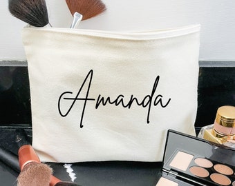 Op de afbeelding: Een witte canvas toilettas met de naam "Amanda" in zwarte cursieve letters. De tas ligt op een zwart oppervlak met make-upkwasten en een palet met oogschaduw.